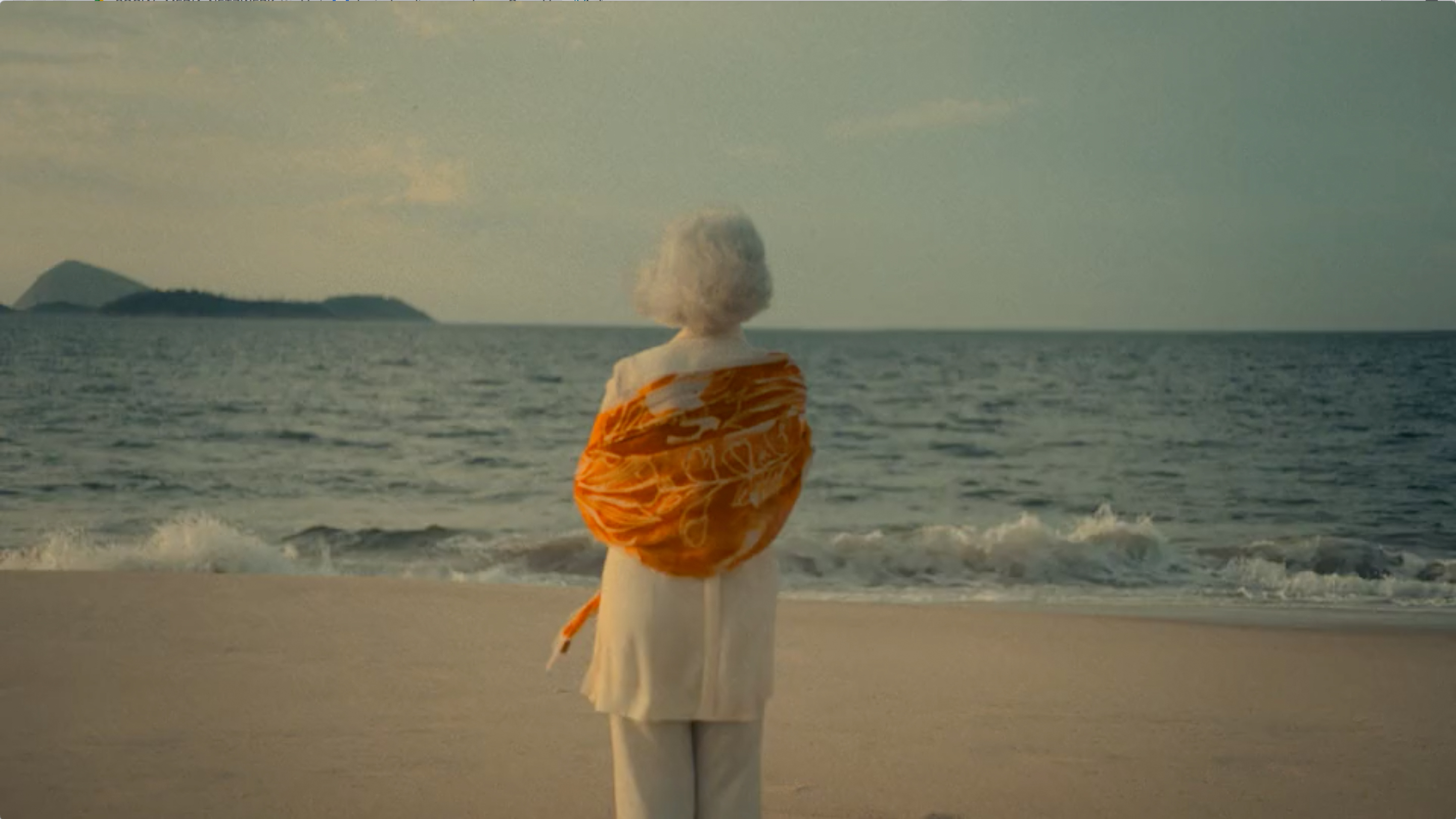 Weißhaarige ältere Dame steht mit dem Rücken zur Kamera in Abendstimmung am Meeresstrand. Am Horizont eine Insel.