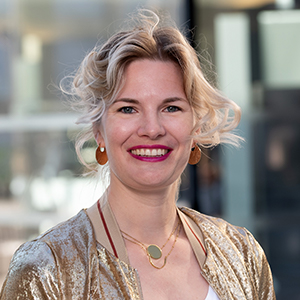 Frau mit blondem Haar, rotem Lippenstift, goldener Jacke, lächelt freundlich