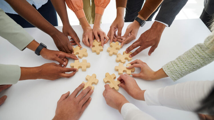 Mehrere Hände fügen gemeinsam große Holzpuzzleteile auf einem Tisch zusammen.