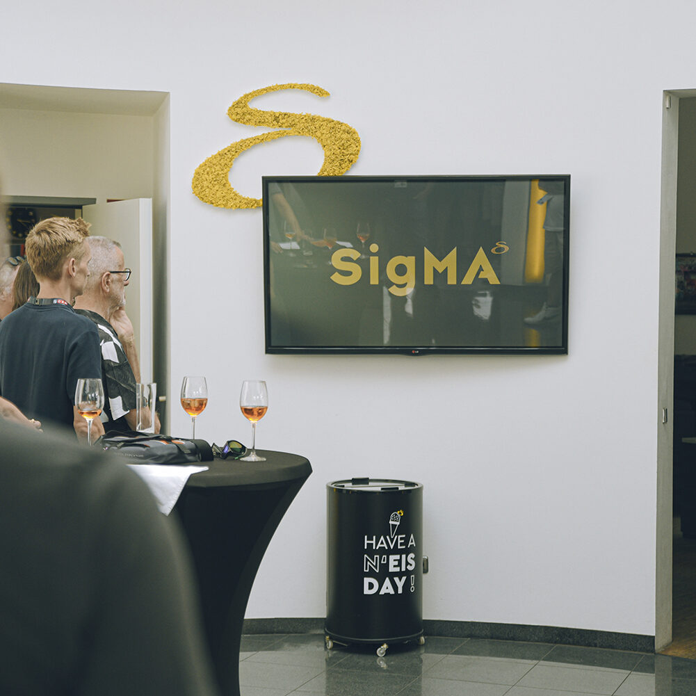 Monitor zeigt SigMA-Logo, Personen hören Präsentation zu