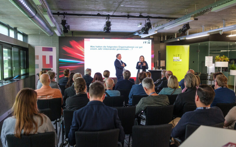 Zwei Vortragender auf der Bühne, davor die Zuhörer in Reihenbestuhlung