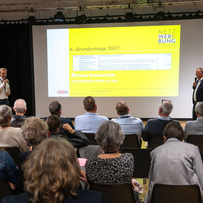 Zwei Vortragende präsentieren vor Publikum auf einer Bühne mit großer Leinwand