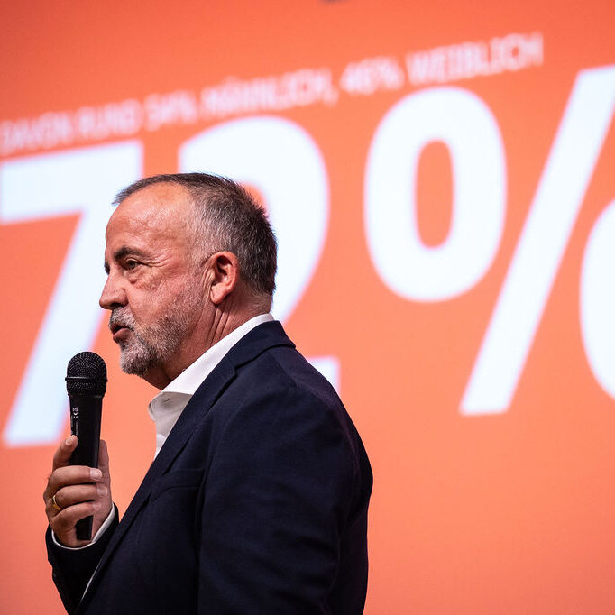 Christoph Schuhmacher spricht in ein Mikrofon, im Hintergrund ein digitaler Screen